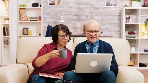 Elderly couple sitting on sofa using laptop for online shopping. Coffee on the table.