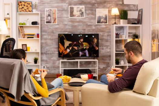 Back view of couple in living room watching a movie on the TV while eating takeaway food