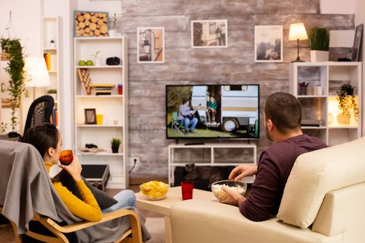 Back view of couple in living room watching a movie on the TV while eating takeaway food