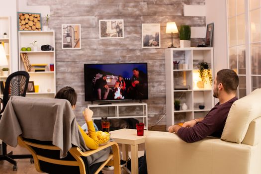 Back view of couple in living room watching a movie on the TV while eating takeaway food