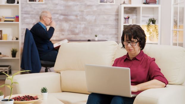 Senior woman with glasses working on laptop sitting on sofa. Elderly age man reading a book in the background.