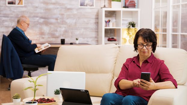 Zoom in shot of elderly age woman with glasses taking a sip of coffee browsing on phone. Elderly age man reading a book in the background.