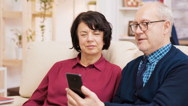 Happy senior couple having a video call seated on couch in their living room. Aged couple using modern technology