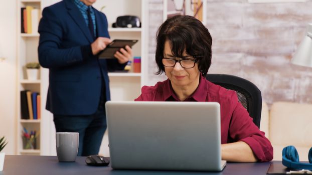 Retired woman working on laptop in living room while her husband is using tablet in the background.