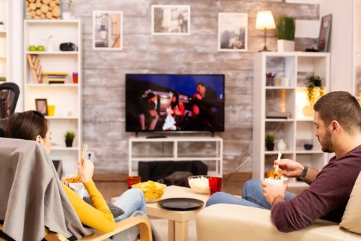 Back view of couple in living room watching a movie on the TV while eating takeaway food