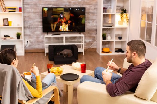 Back view of couple in living room watching a movie on the TV while eating takeaway food