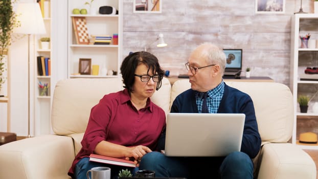 Elderly couple sitting on sofa using laptop for online shopping. Coffee on the table.