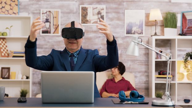 Senior man using virtual reality goggles in living room. Woman watching tv while sitting on the sofa in the background.