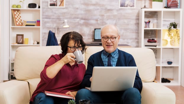 Elderly couple sitting on sofa using laptop for online shopping. Coffee on the table.