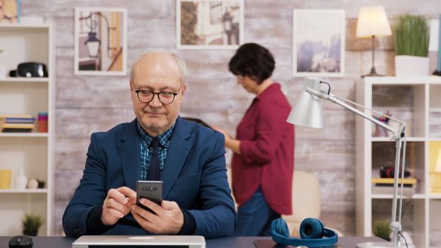 Elderly man with glasses browsing on mobile phone in living room. Woman walking and using tablet in the background.