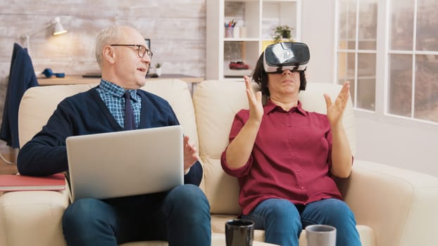 Amazed senior woman while using virtual reality goggles on sofa with her husband next to her using laptop.