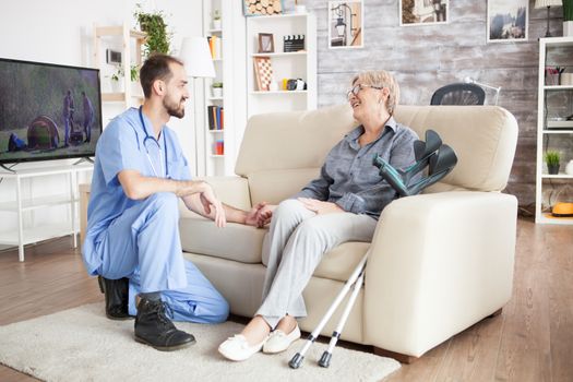 Joyful old woman in a nursing home talking with her health visitor while sitting on the couch with crutches next to her.