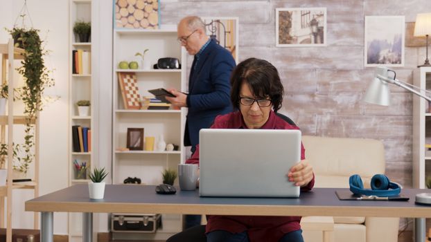 Elderly woman opening laptop and putting his glasses on her face. Old man with glasses using tablet in the brackground.