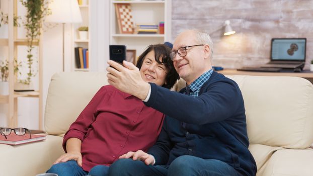 Happy senior couple sitting on sofa taking a selfie in the living room