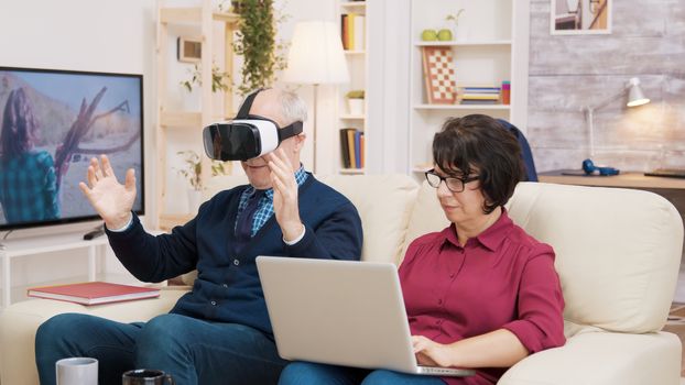 Elderly age woman sitting on sofa using laptop while her husband is experiencing virtual reality for the first time