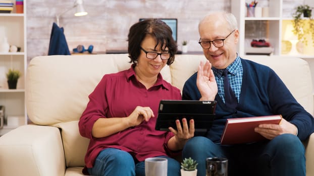 Senior couple waving at tablet during a video call. Elderly people sitting on sofa.