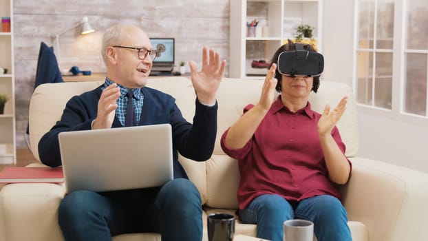 Amazed senior woman while using virtual reality goggles on sofa with her husband next to her using laptop.