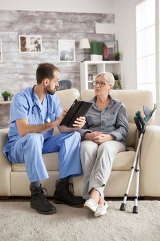 Old woman with crutches in nursing home sitting on couch listening male doctor with tablet computer.