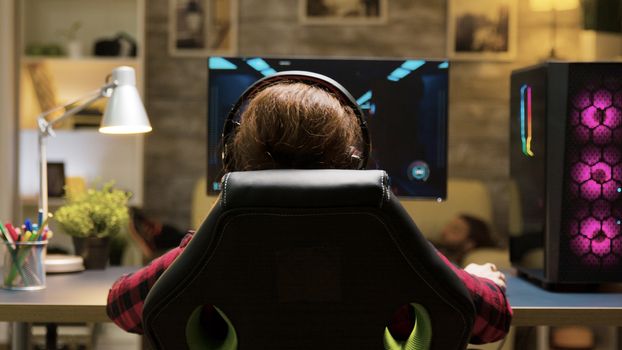 Dolly shot of woman sitting on gaming chair playing computer games. Man lying on sofa in the background.