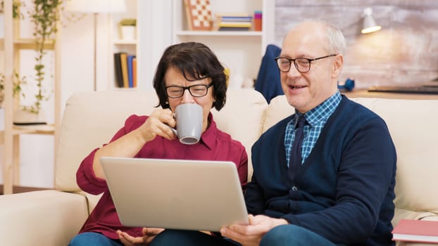 Elderly age couple sitting on sofa holding laptop during a video call. Couple waving at laptop.