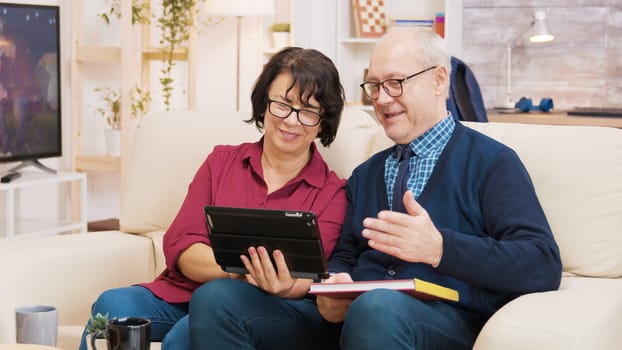 Old couple sitting on sofa during a video call on tablet. Old couple waving at tablet.