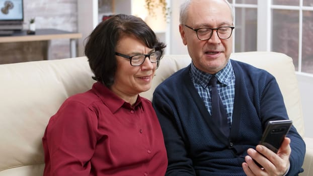 Happy elderly couple sitting on sofa holding smartphone during a video call.