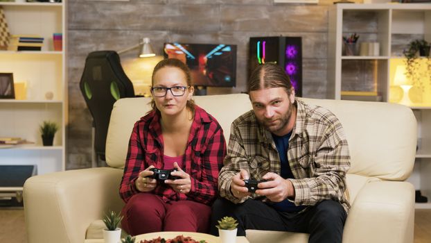 Excited couple giving high five while playing online video games sitting on couch using wireless controllers.