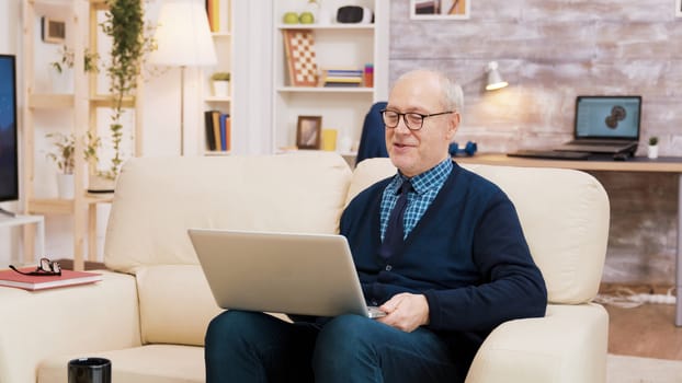 Elderly age couple with glasses sitting on sofa during a video call on laptop. Aged couple using modern technology