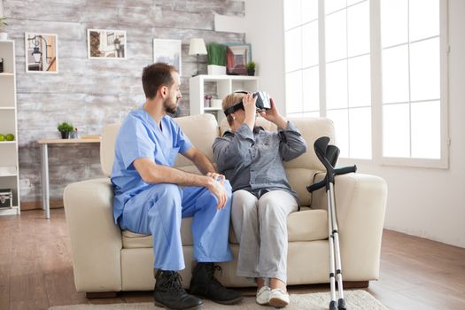 Bearded doctor sitting on couch in nursing home while old woman with crutches uses virtual reality glasses.