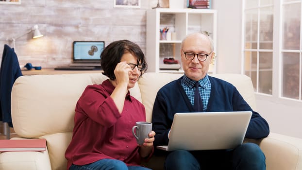 Grandparents with glasses sitting on sofa talking with family during a video call on laptop.
