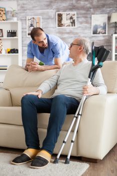 Senior man sitting on couch with crutches in nursing home talking with male nurse.