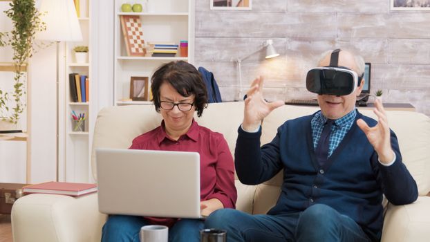 Senior man trying a VR headset in the living room while his wife uses a laptop next to him. Modern old couple using technology