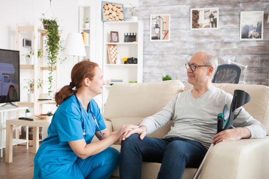 Happy old man with crutches sitting down on couch in nursing home talking with female doctor.