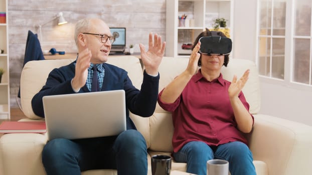 Amazed senior woman while using virtual reality goggles on sofa with her husband next to her using laptop.