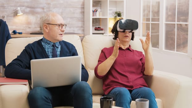 Amazed senior woman while using virtual reality goggles on sofa with her husband next to her using laptop.