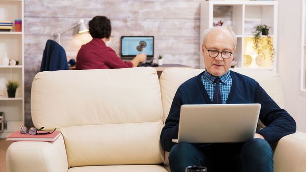 Happy senior middle aged couple sitting on sofa waving at laptop during a video call.