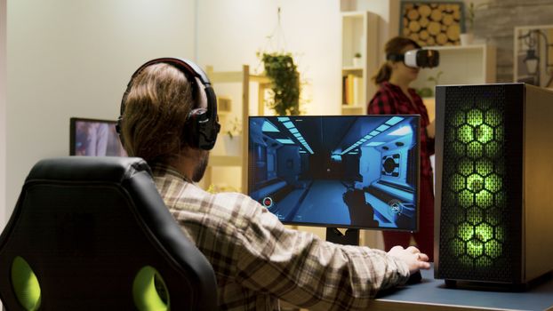Man putting headphones on and start playing video games on computer. Woman experiencing virtual reality with vr headset in the background.
