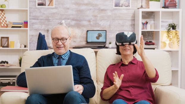 Elderly age woman sitting on sofa wearing virtual reality goggles. Old man sitting on sofa using laptop next to his wife