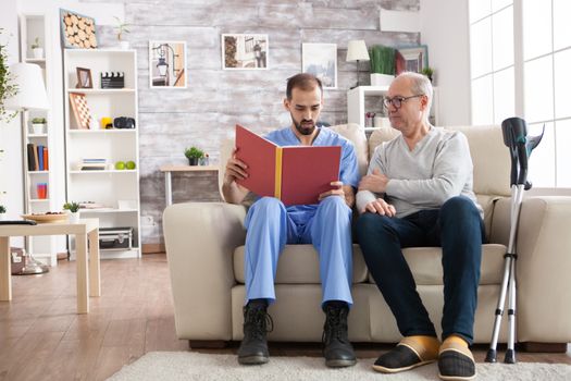 Bearded doctor reading a story in nursing home for an old man. Elderly age man with crutches.