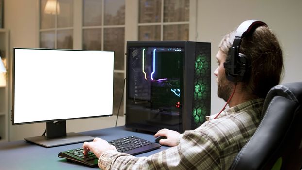 Man with headphones sitting on gaming chair playing games on computer with green screen.