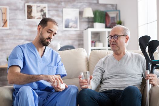 Caucasian young doctor in nursing home giving pills to senior man with crutches. .