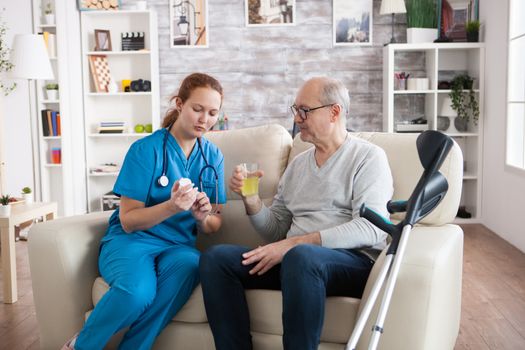 Lady caregiver giving senior man with crutches pills for his disease in nursing home.