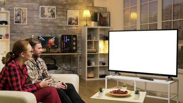 Relaxed couple sitting on sofa playing video games using wireless joysticks on tv with green screen.