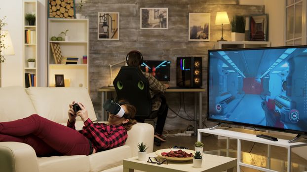 Woman lying on sofa playing video games using vr headset in living room. Boyfriend playing on computer.