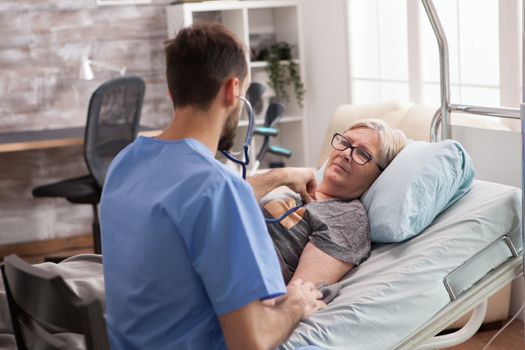 Old woman in nursing home looking at male doctor while he is checking herheart beat using stethoscope.