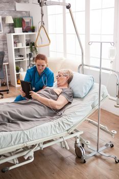 Female doctor helping old woman in nursing home to use wifi on tablet computer.