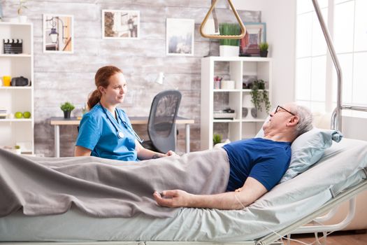 Female nurse talking with old man in nursing home sitting on laying on bed.