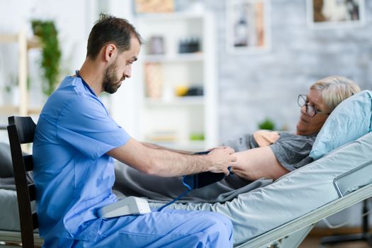 Bearded health visitor in a nursing home with stethoscope checking old lady blood pressure using digital device.