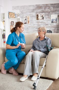 Female nurse sitting on couch with senior woman telling her how to take her pills in nursing home.