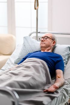 Retired old man in nursing home laying on bed with transfusion attached to his arm